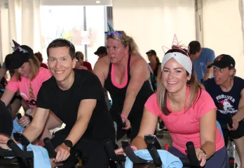 A group of people in pink and black shirts cycle on stationery bikes at a SWEAT for Strong Kids event.