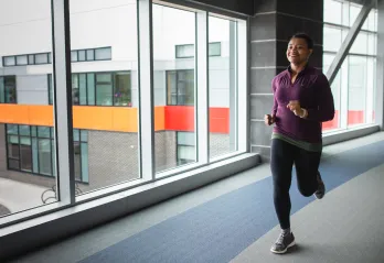 A woman in a purple shirt and black leggings jogs on an indoor walking track.