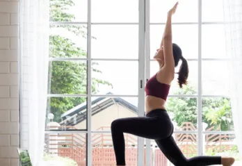 A woman practices yoga by a window at home, following an online workout on her laptop.