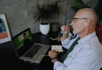 A man in a white button-up shirt with glasses and headphones sits at a computer desk while having a video call with another man on his laptop.