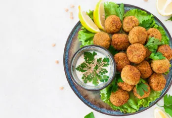 A blue bowl of falafels on a bed of lettuce with a side sauce.