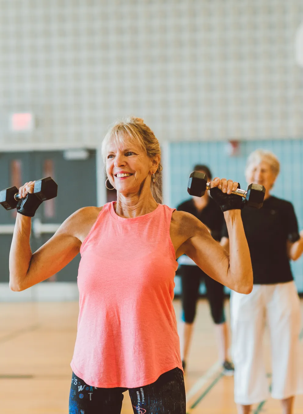 An older woman in a pink tank top smiles and lifts weights as part of a group fitness class.