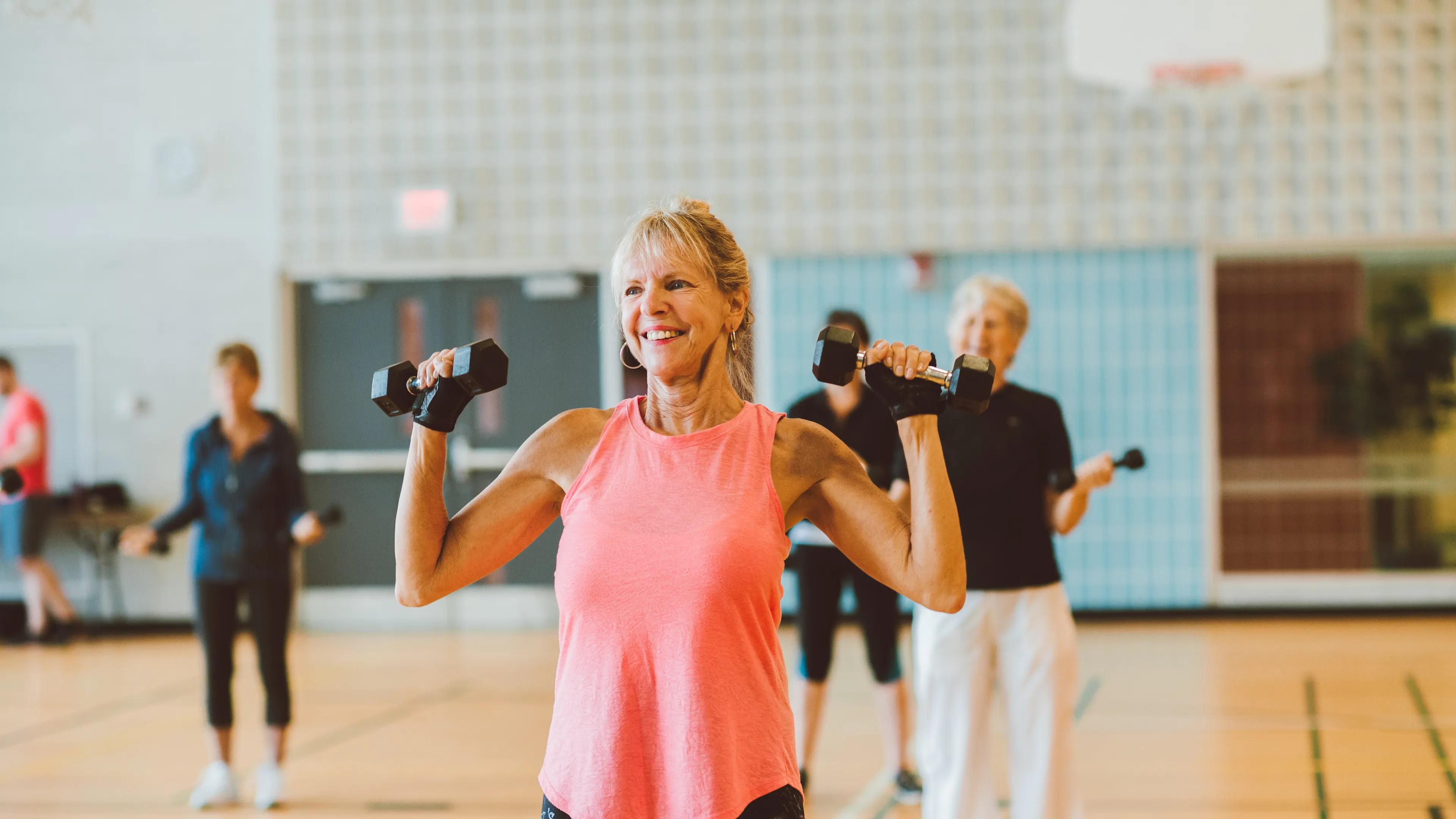 An older woman in a pink tank top smiles and lifts weights as part of a group fitness class.