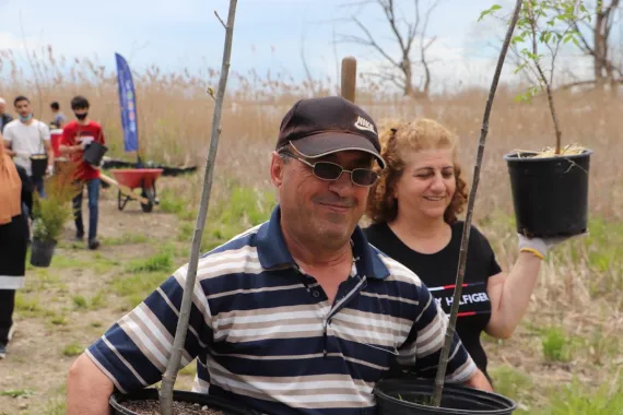 A man in a striped shirt, sunglasses and hat, carries trees with a smiling woman in a black t-shirt at a tree planting event.