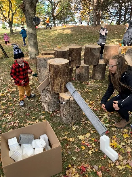 An educator observes a young boy in a red plaid jacket put together loose parts from a box outside.