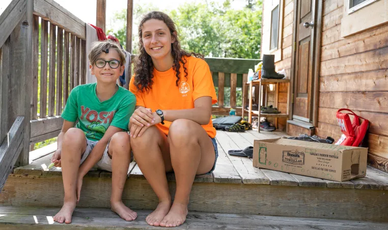 A counsellor and camper smile and sit together on a wooden deck.