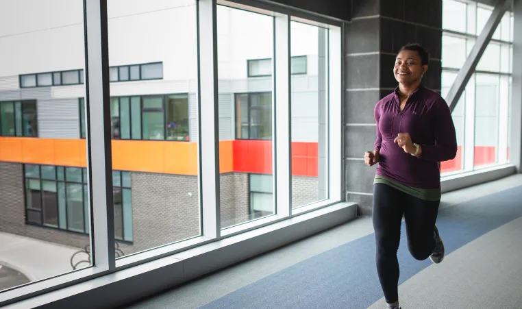 A woman in a purple shirt and black leggings jogs on an indoor walking track.