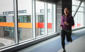 A woman in a purple shirt and black leggings jogs on an indoor walking track.