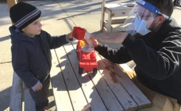 A young boy and Early Childhood Educator in PPE play at an outdoor picnic table with a red truck.