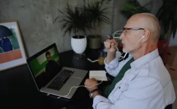 A man in a white button-up shirt with glasses and headphones sits at a computer desk while having a video call with another man on his laptop.